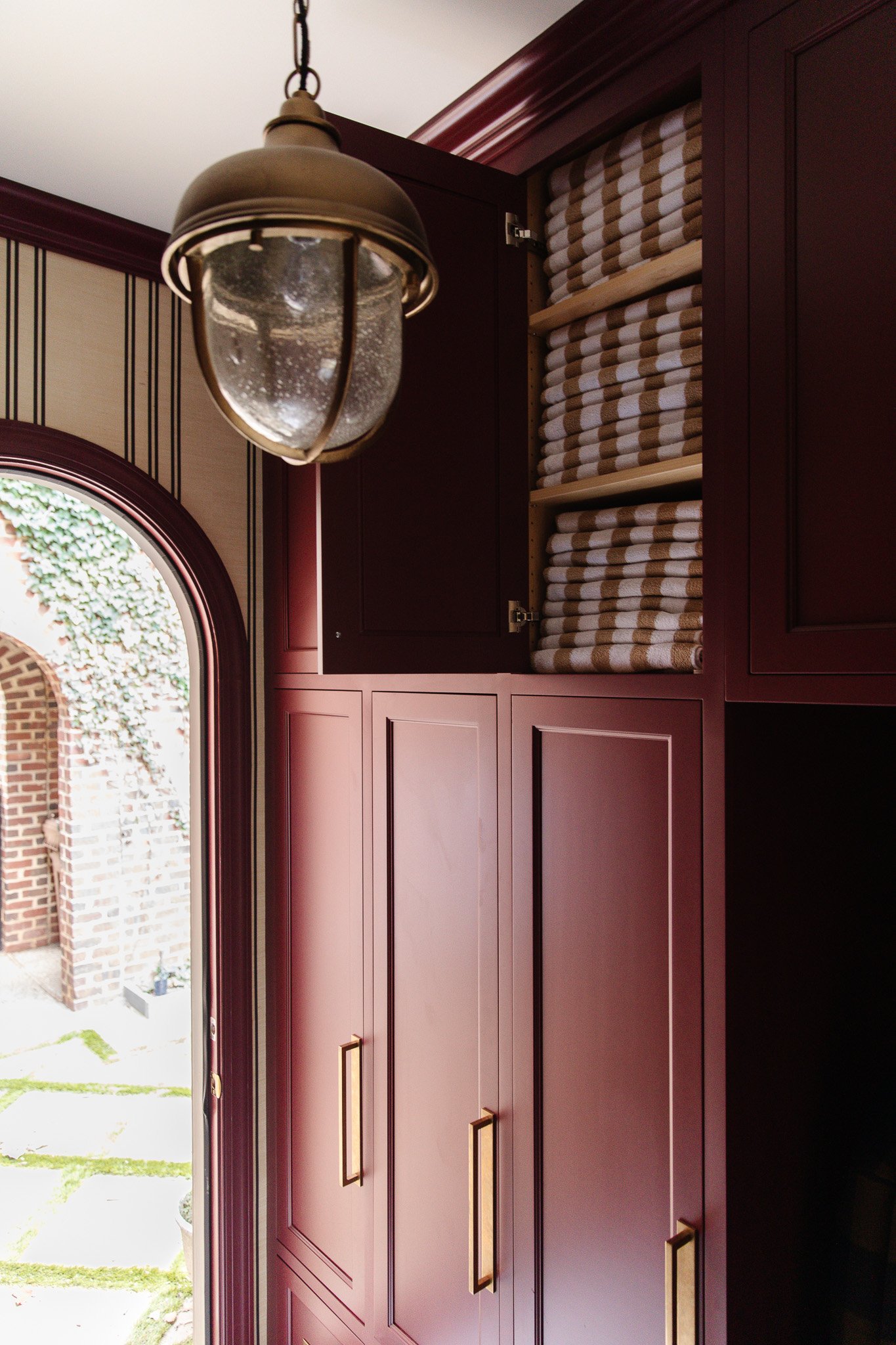 Chris Loves Julia | Moody dark red mudroom with an upper cabinet open to reveal a stack of white and beige pool towels