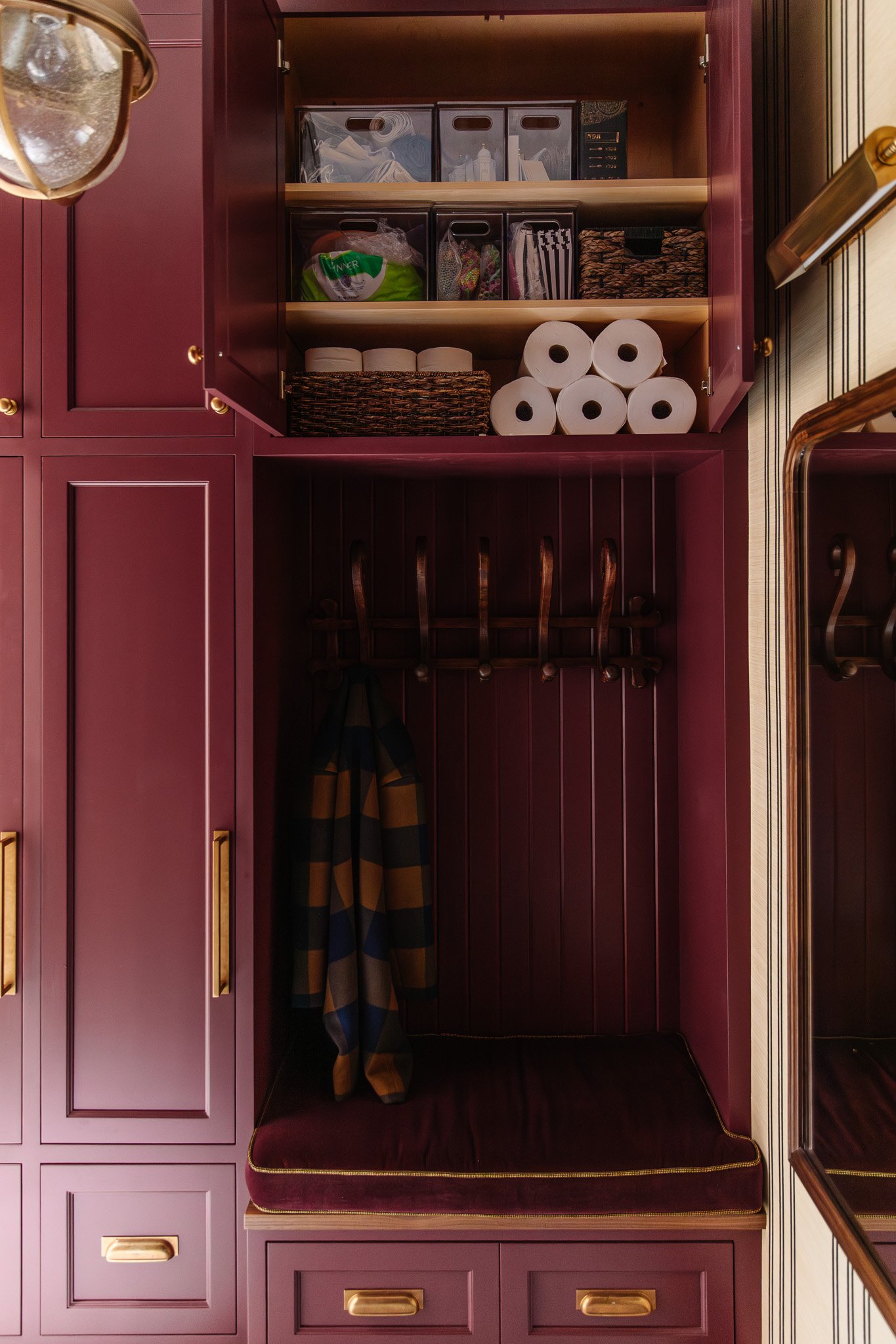 Chris Loves Julia | Moody dark red mudroom with upper cabinets open to reveal storage bins and paper towels