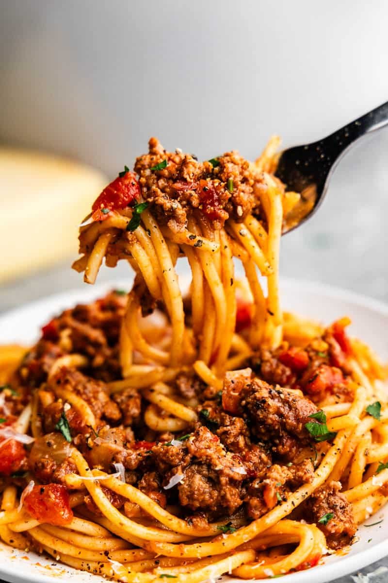 A close up of a fork lifting a bite of spaghetti noodles with meaty sauce from a plate.