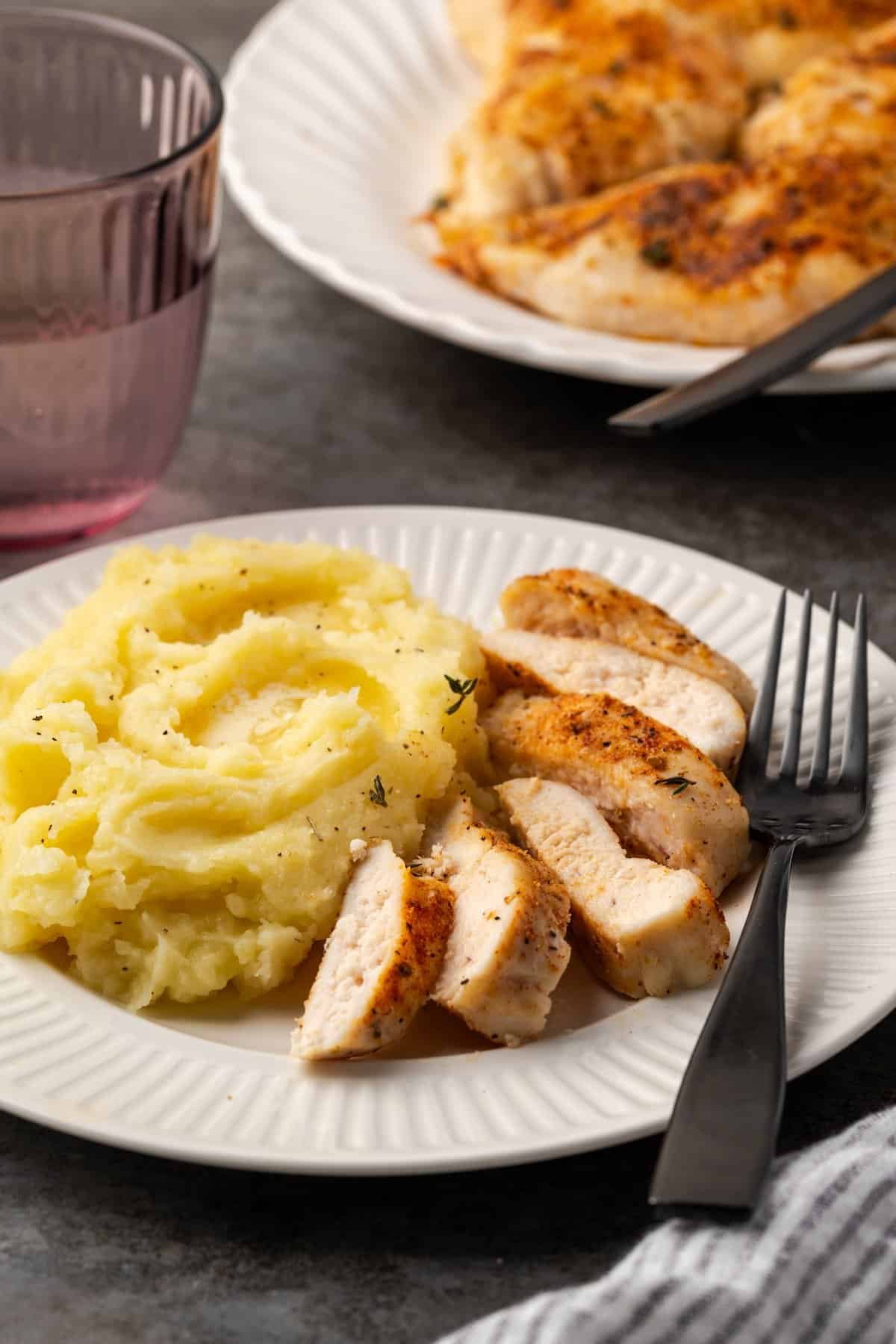 Baked chicken breast cut into slices next to a serving of mashed potatoes and a fork on a white plate, with a platter of chicken in the background.