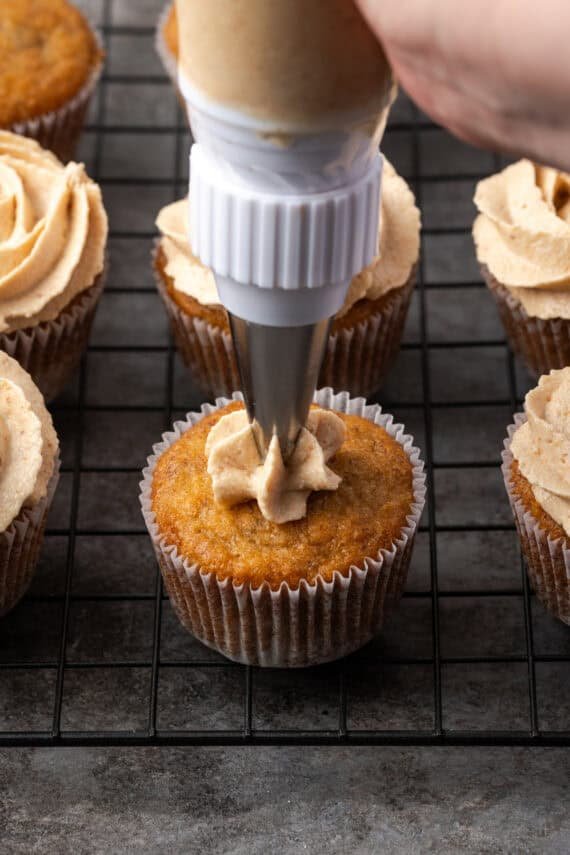 A piping tip pipes frosting onto a banana cupcake on a wire rack, surrounded by more frosted cupcakes.