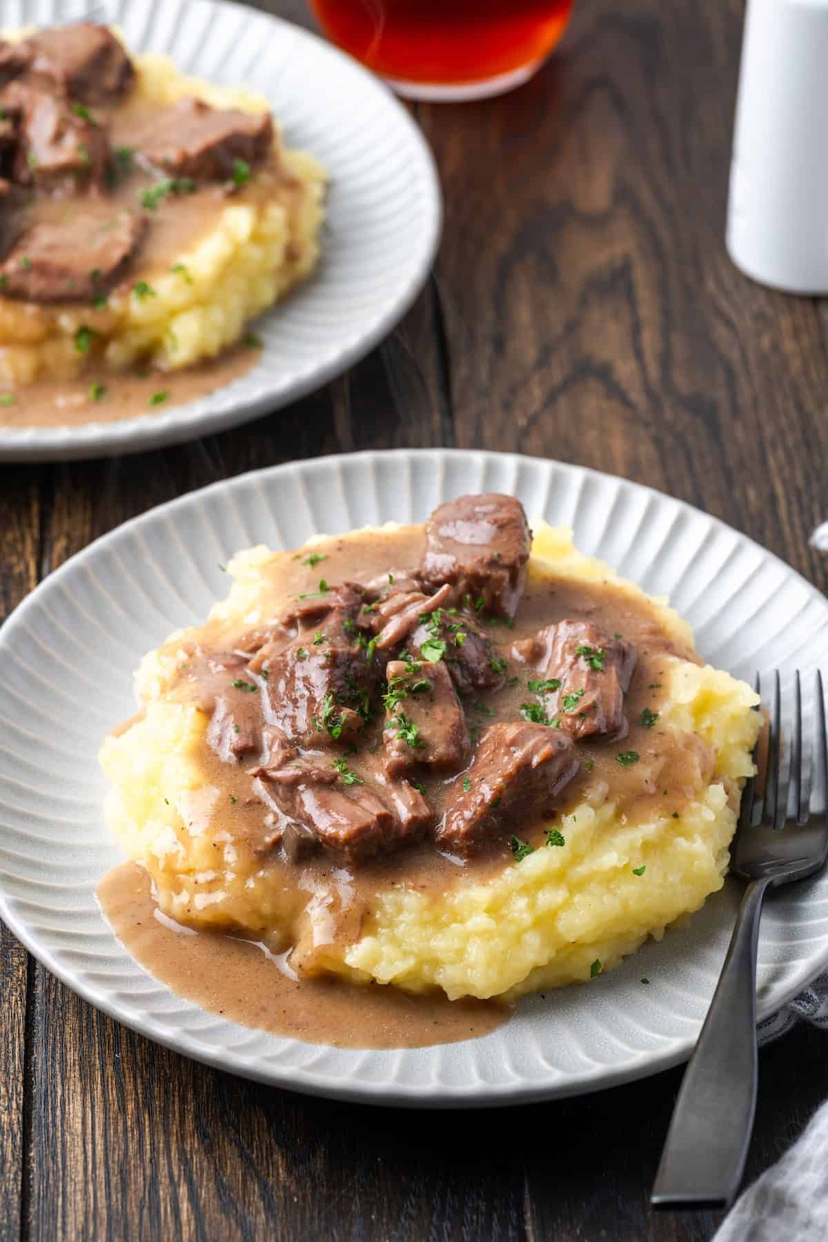 No peek beef tips and gravy served over mashed potatoes on a white plate next to a fork, with a second plate of beef tips in the background.