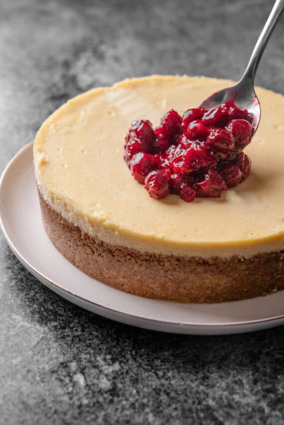 Cherry pie filling being spooned over top of a baked cheesecake on a large plate.