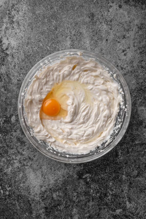 An egg added to cheesecake batter in a glass mixing bowl.