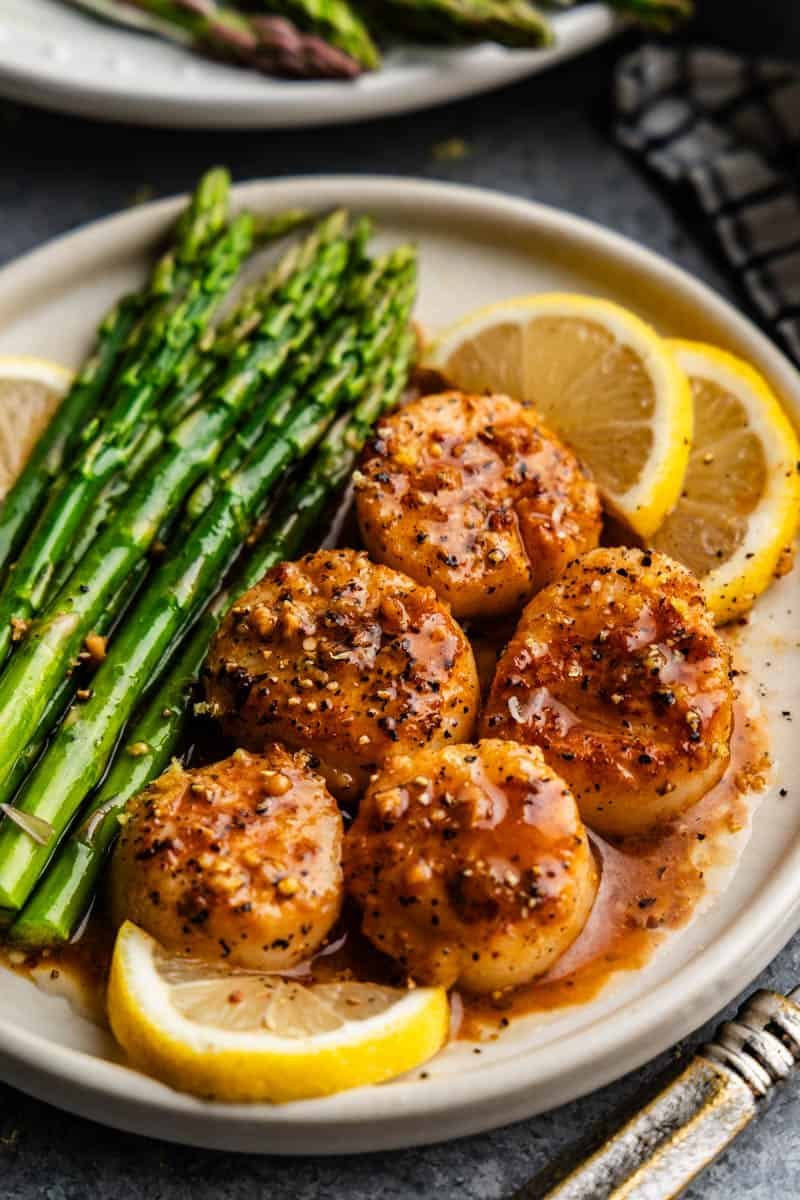 A plate of lemon garlic scallops with lemon slices and asparagus on the side.